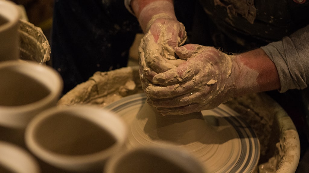 Hands shaping a clay vessel on a potter's wheel. Ceramic cups are out-of-focus in the foreground.